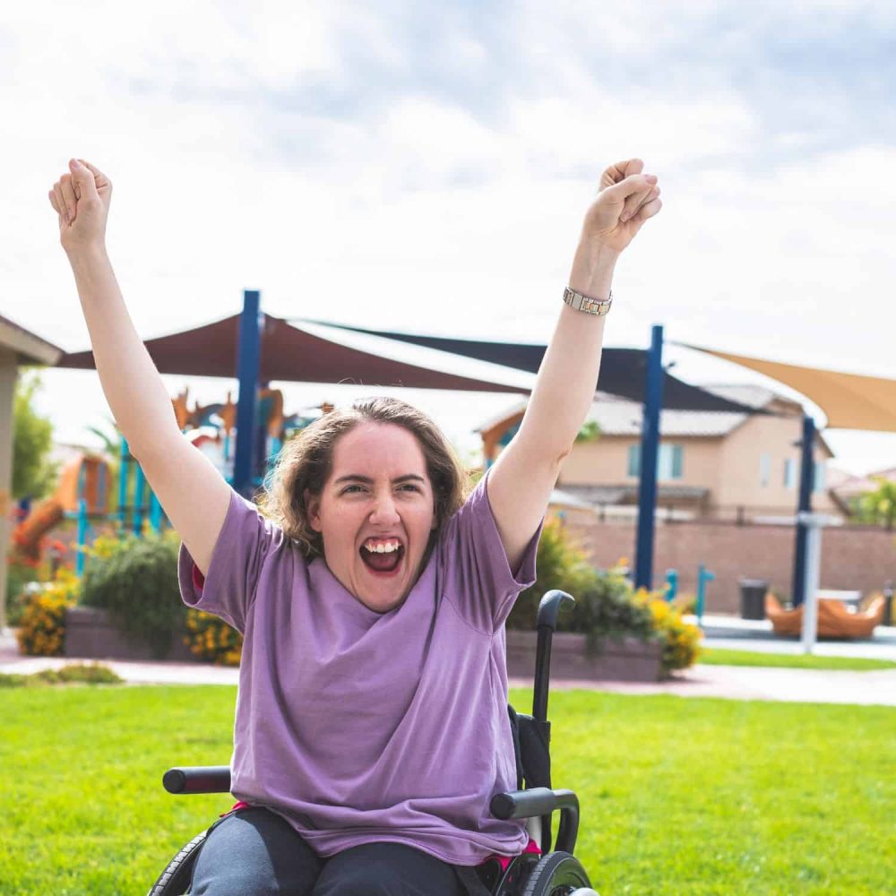 Woman in wheelchair celebrating outdoors with arms raised and smiling.